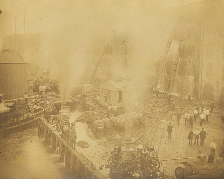 Firefighting scene showing dockside building on fire, being put out by hose and ladder apparatus of four fire engines, with spectators