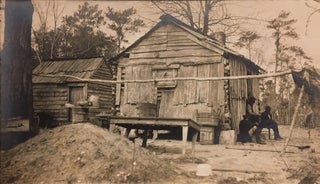 Item #314042 Photograph of three black men sitting outside clapboard-covered log cabin and shed
